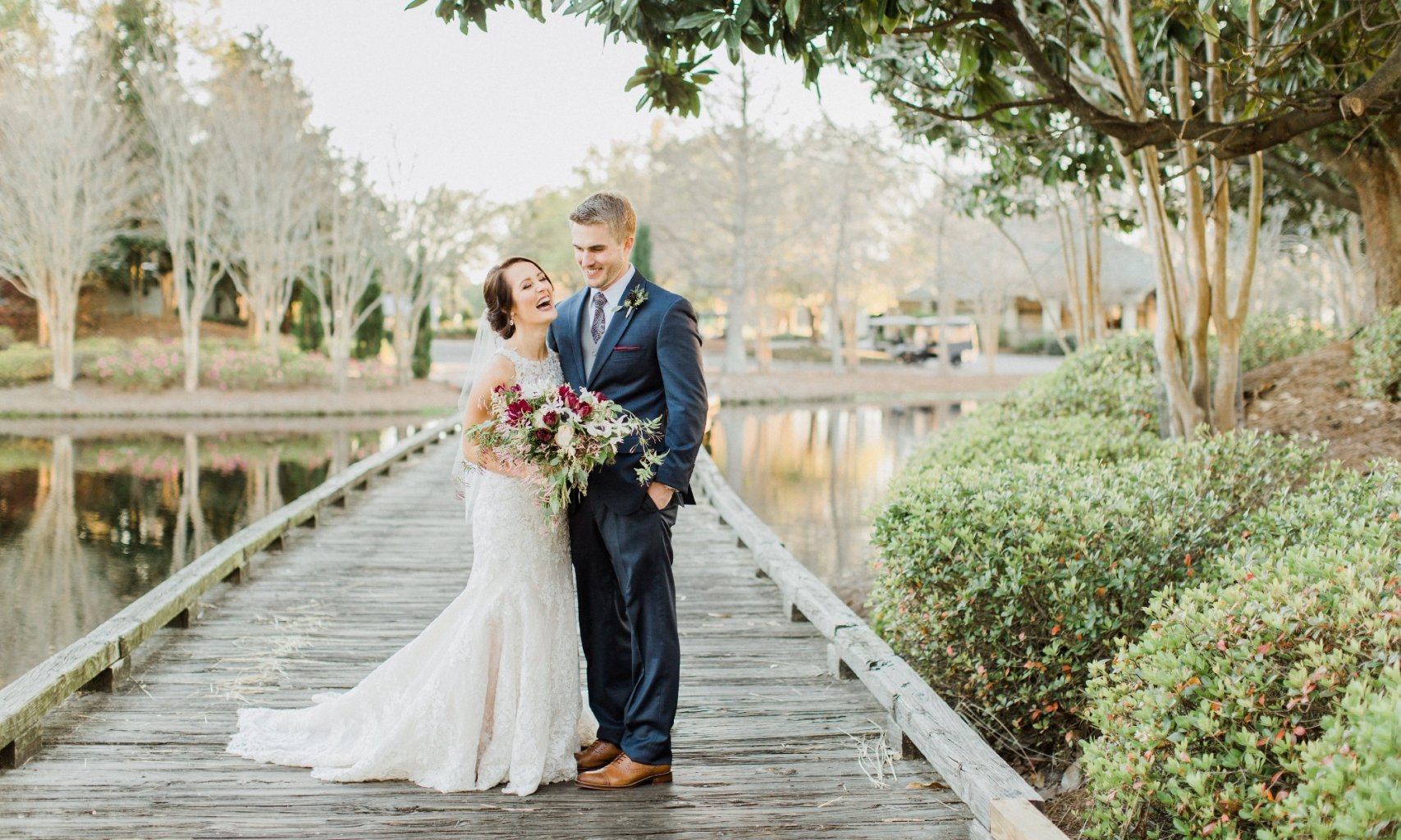 A laughing bride and groom laughing on a wooden bridge with a pnd in the background