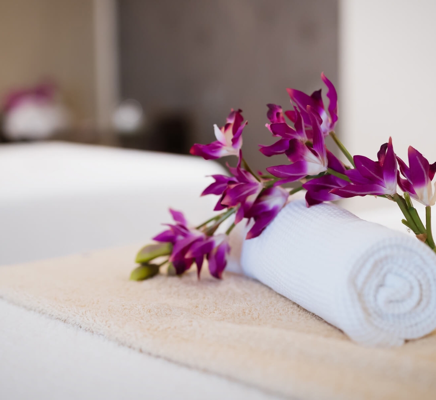 A white towel rolled up and placed on a bed with a flower laying beside it